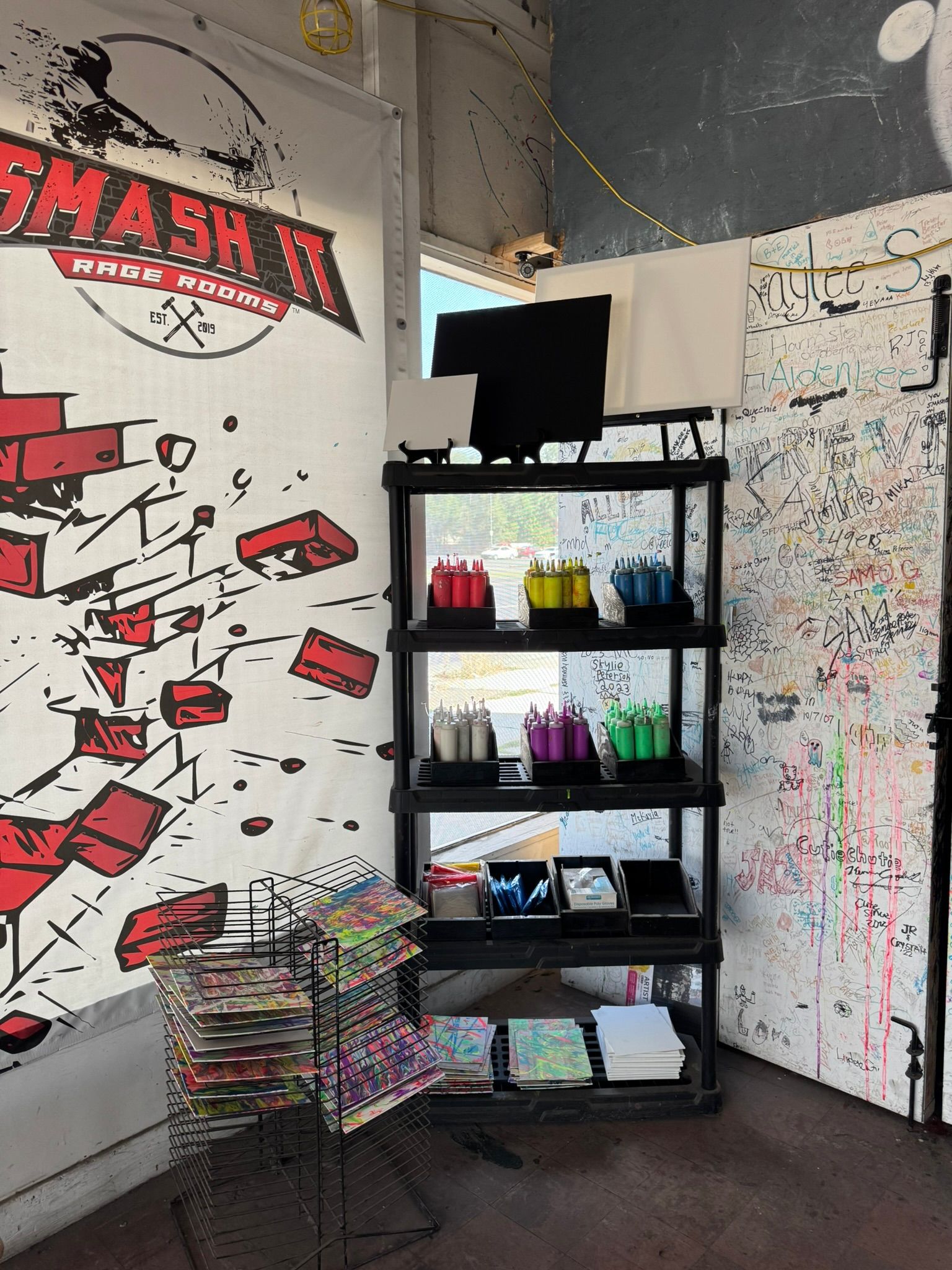 Vibrant indoor art station in a studio corner with black shelving holding organized squeeze bottles of paint (red, yellow, blue, purple, green), blank canvases, paint trays, stacks of colorful panels on drying racks and a graffiti-covered signature wall.