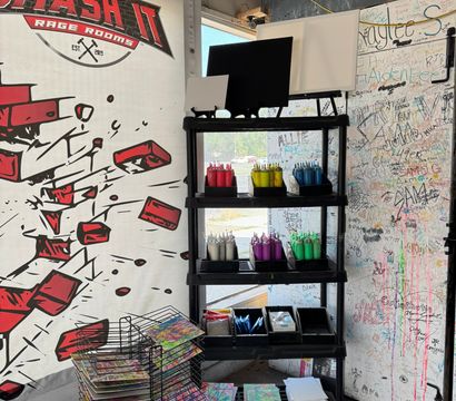 Vibrant indoor art station in a studio corner with black shelving holding organized squeeze bottles of paint (red, yellow, blue, purple, green), blank canvases, paint trays, stacks of colorful panels on drying racks and a graffiti-covered signature wall.