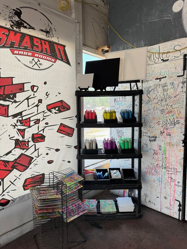 Vibrant indoor art station in a studio corner with black shelving holding organized squeeze bottles of paint (red, yellow, blue, purple, green), blank canvases, paint trays, stacks of colorful panels on drying racks and a graffiti-covered signature wall.