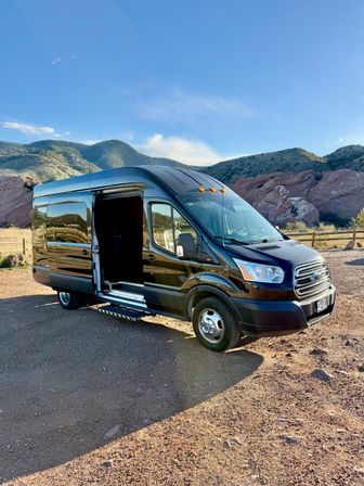Black camper van with open sliding door parked on a dirt turnout against red rock formations and tree-covered hills under a bright blue sky — scenic mountain camping scene.