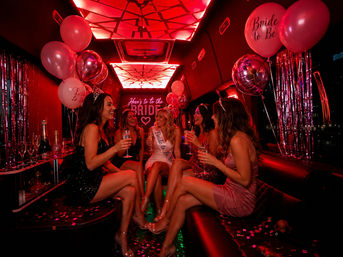 Group of women celebrating a bachelorette party inside a pink-red neon-lit party bus, bride-to-be wearing sash and tiara, holding champagne with balloons, confetti and festive decor.