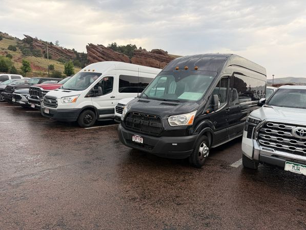 Fleet of black and white passenger vans and pickup trucks parked on wet asphalt with red sandstone cliffs rising behind under a cloudy Colorado sky.