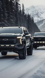 Two matte-black off-road pickup trucks with oversized snow tires and LED light bars driving on a snowy mountain road through evergreen forest, misty alpine peaks in the background