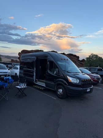 Black camper van with open side door parked at sunset in a red‑rock parking lot, folding chairs and a small table with drinks in the foreground and pastel clouds overhead.