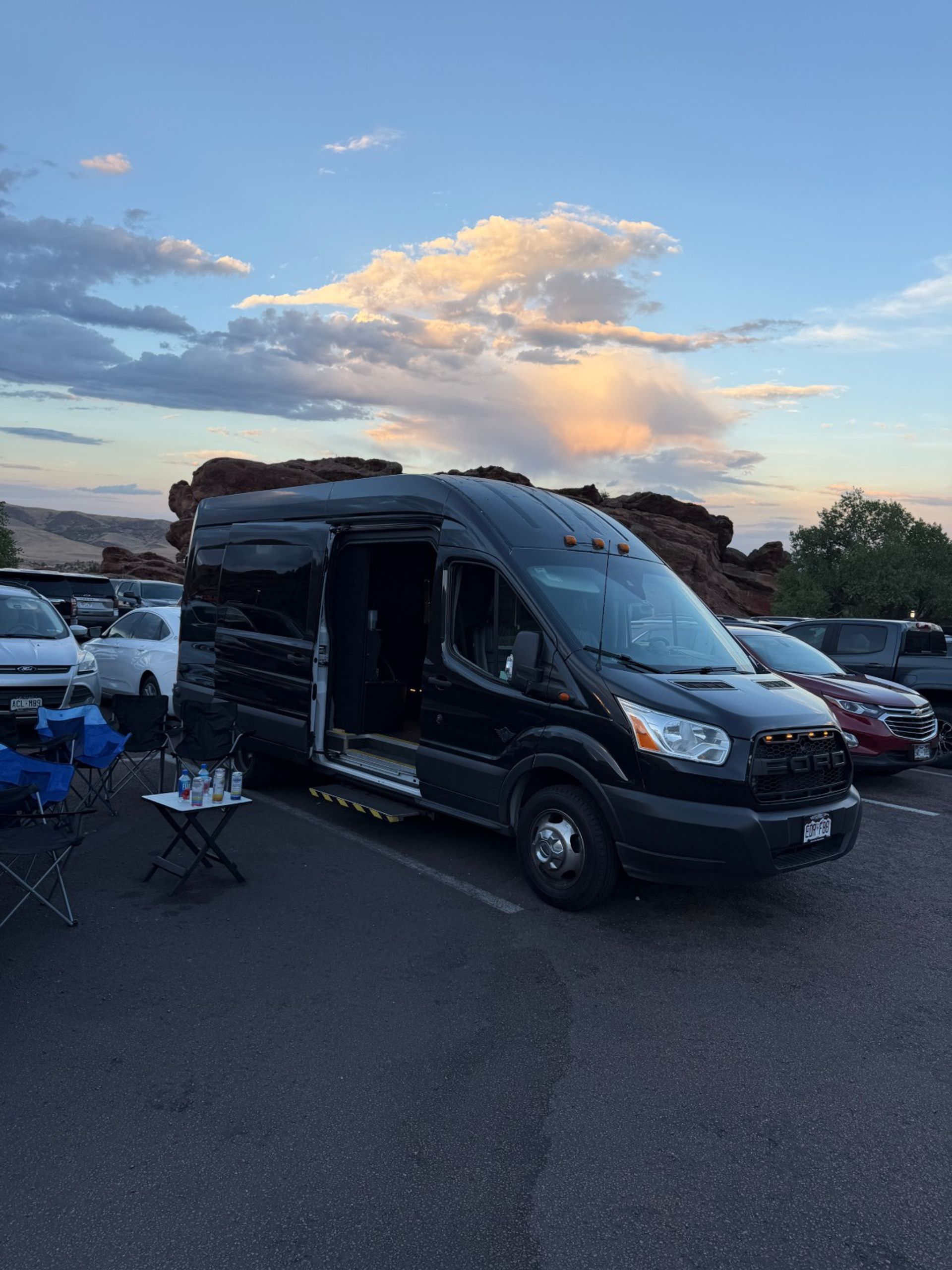 Black camper van with open side door parked at sunset in a red‑rock parking lot, folding chairs and a small table with drinks in the foreground and pastel clouds overhead.