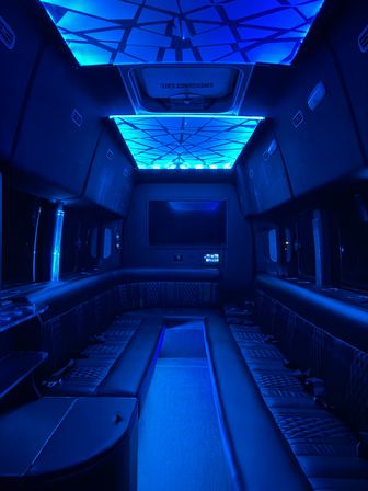 Spacious party bus interior with wraparound leather benches, geometric blue LED ceiling panels, flatscreen monitor and cup holders, mood-lit luxury shuttle cabin