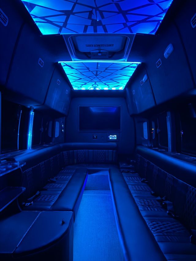 Spacious party bus interior with wraparound leather benches, geometric blue LED ceiling panels, flatscreen monitor and cup holders, mood-lit luxury shuttle cabin