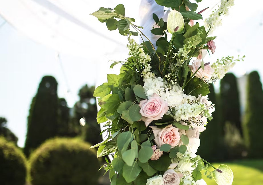 Romantic outdoor wedding floral arrangement of soft pink roses, white blooms and eucalyptus greenery wrapped around an arch post against a sunlit garden with blurred hedges.
