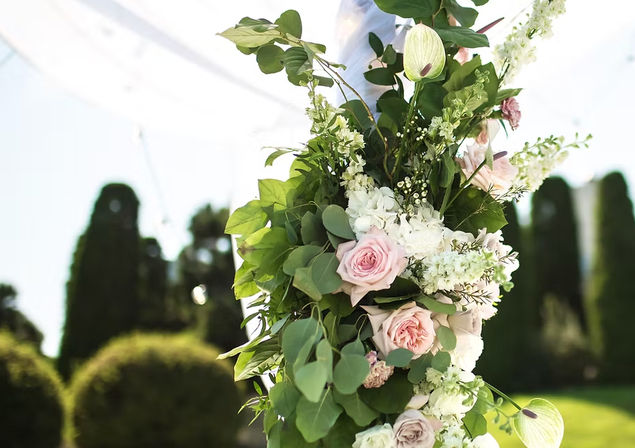 Romantic outdoor wedding floral arrangement of soft pink roses, white blooms and eucalyptus greenery wrapped around an arch post against a sunlit garden with blurred hedges.