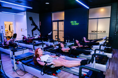 Row of participants in a reformer Pilates class in a modern neon-lit fitness studio doing synchronized leg lifts and core work on reformer machines.