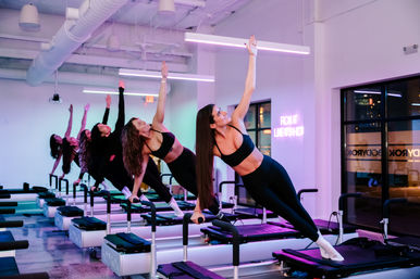 Group of women in athletic wear doing side-plank reformer Pilates on machines in a sleek, neon-lit urban fitness studio