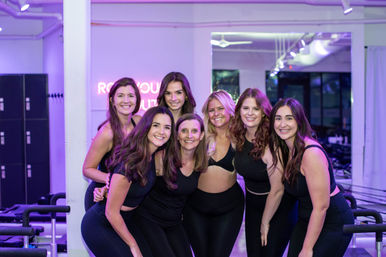 Seven smiling women in black activewear posing together in a purple-lit pilates reformer studio with machines and lockers in the background