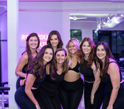 Seven smiling women in black activewear posing together in a purple-lit pilates reformer studio with machines and lockers in the background