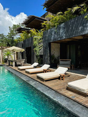 Turquoise pool and wooden deck at a modern tropical villa — cushioned sun loungers, black concrete facades, bamboo pergolas and lush palm greenery
