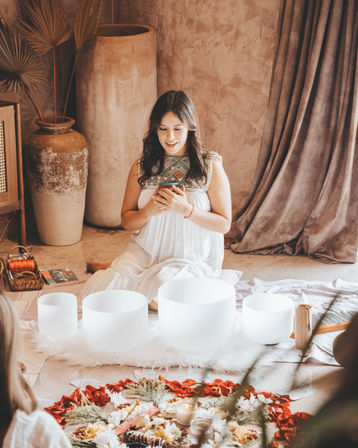 Woman in a white embroidered dress seated in a boho studio holding a phone, surrounded by white crystal singing bowls on a fluffy rug, a flower-and-petal mandala with candles on the floor, and large clay vases and draped curtains in the background — intimate sound-bath meditation scene.