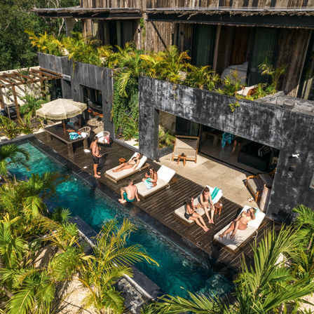 Aerial view of a tropical resort pool deck with sun loungers and sunbathers, a narrow turquoise pool, weathered concrete balconies topped with palm planters and lush greenery.