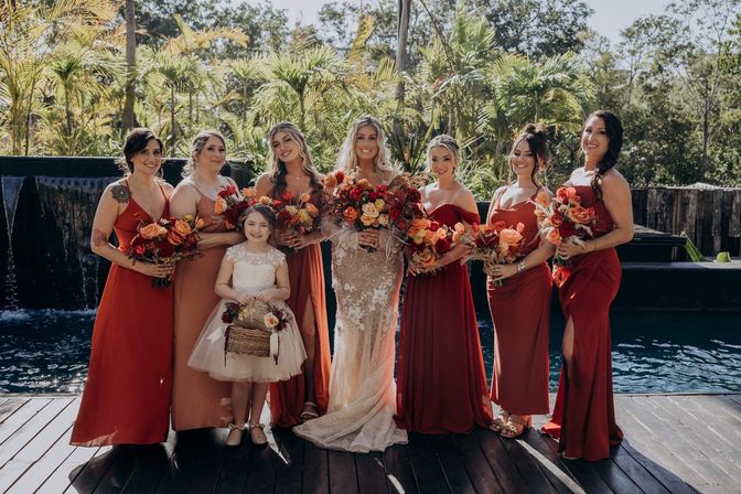 Bride in lace gown with bridesmaids in red and rust dresses and a flower girl holding a basket, all holding orange and red bouquets on a sunny tropical poolside deck with palm trees — outdoor wedding portrait.
