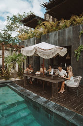 Group of friends dining under a fringed macramé umbrella at a wooden table beside a turquoise-tiled pool at a tropical villa with lush palms and modern concrete architecture