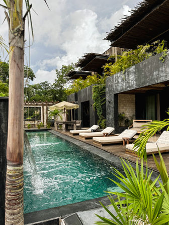 Tropical boutique poolside scene — turquoise lap pool with bamboo waterfall, wooden deck and cushioned sun loungers, lush palms and overhanging thatched balconies at an eco-luxury retreat.