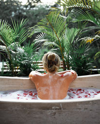Back view of a person relaxing in a rose-petal bubble bath at a tropical outdoor spa, framed by lush palm plants for a serene wellness escape