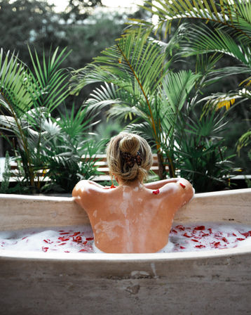 Back view of a person relaxing in a rose-petal bubble bath at a tropical outdoor spa, framed by lush palm plants for a serene wellness escape