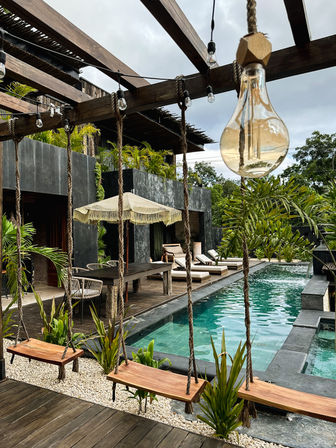 Tropical modern poolside patio with wooden swings, hanging Edison bulbs on a rope pergola, lounge chairs and a turquoise tiled pool framed by lush greenery.