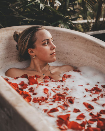 Woman relaxing in a bubble bath filled with red rose petals in a stone tub, gazing sideways with tropical plants in the background for a serene outdoor spa vibe