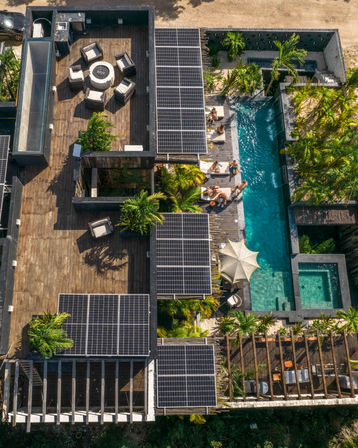 Aerial view of a tropical villa rooftop with solar panels, wooden deck seating around a circular fire pit, palm trees and a turquoise swimming pool with loungers and people relaxing.