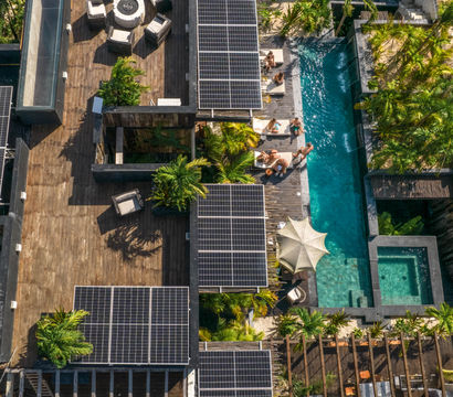 Aerial view of a tropical villa rooftop with solar panels, wooden deck seating around a circular fire pit, palm trees and a turquoise swimming pool with loungers and people relaxing.