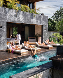 Tropical outdoor pool deck at a modern villa with sunbathers on wooden chaise lounges and a person mid-dive splashing into a turquoise pool beneath a concrete balcony with potted plants