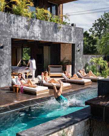 Tropical outdoor pool deck at a modern villa with sunbathers on wooden chaise lounges and a person mid-dive splashing into a turquoise pool beneath a concrete balcony with potted plants