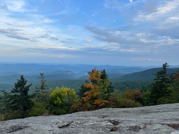 Panoramic mountain overlook with layered blue ridgelines, rocky foreground and early autumn trees beneath a wide sky