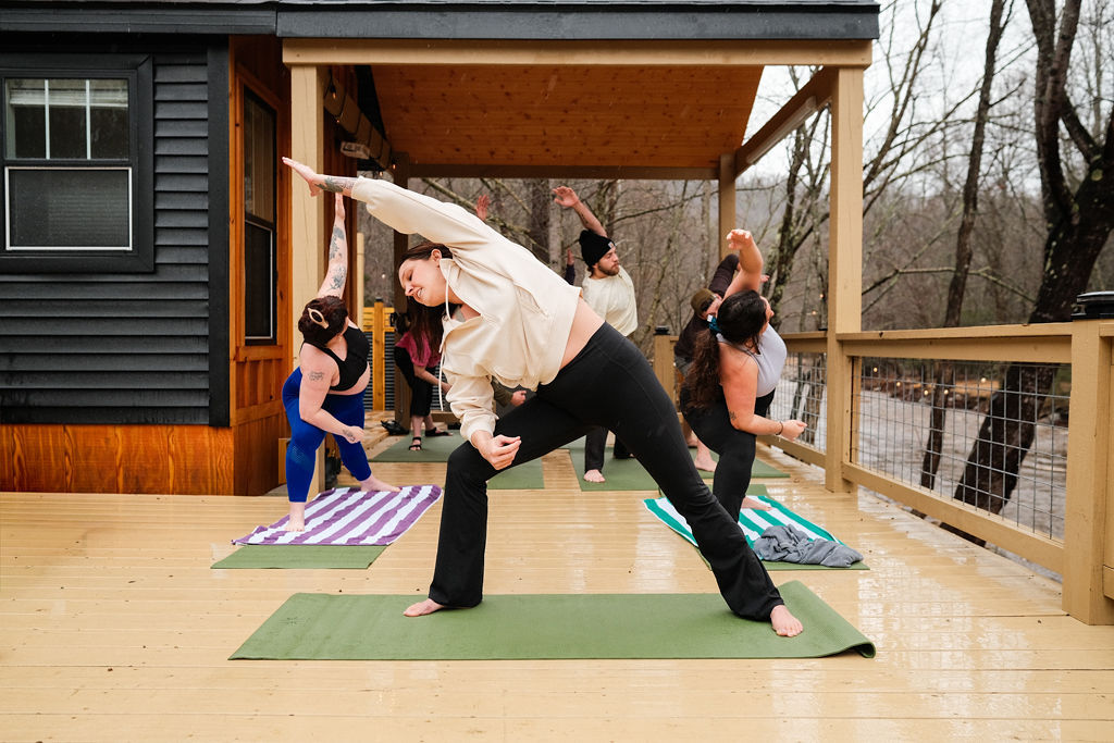 Small group yoga class on an outdoor wooden deck overlooking a riverside forest, participants practicing side-angle stretches on mats under a covered porch on a wet, overcast day.