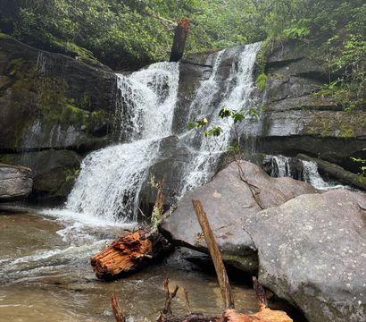 Tiered waterfall cascading over moss-covered boulders into a shallow pool with orange fallen logs in a lush green forest