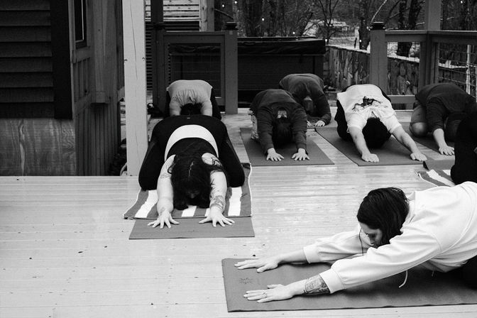 Black-and-white shot of an outdoor yoga class on a wooden deck, several people on mats stretching forward in child’s pose beside a railing and trees.