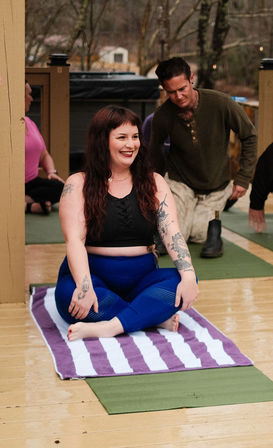 Smiling woman in blue leggings and black top sitting cross-legged on a purple-and-white striped towel during an outdoor group yoga class on a wooden deck