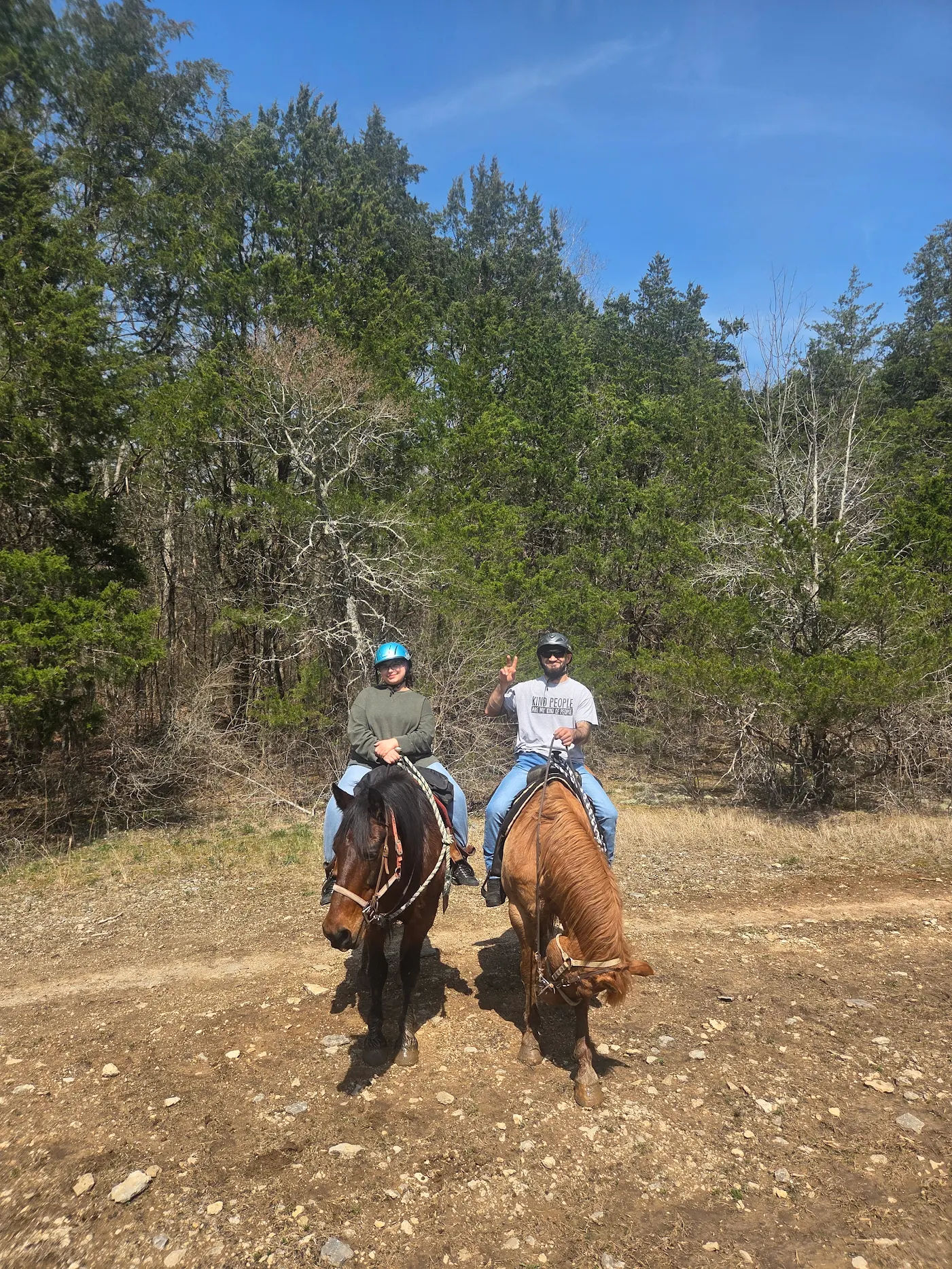 Two helmeted riders on brown horses on a sunny dirt trail through a wooded forest with blue sky, one rider flashing a peace sign.