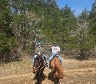 Two helmeted riders on brown horses on a sunny dirt trail through a wooded forest with blue sky, one rider flashing a peace sign.