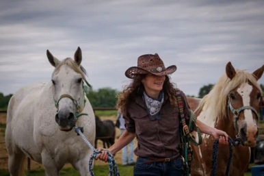 Cowgirl in a brown cowboy hat and bandana leading a white horse and a brown-and-white horse across a grassy ranch pasture under a cloudy sky.
