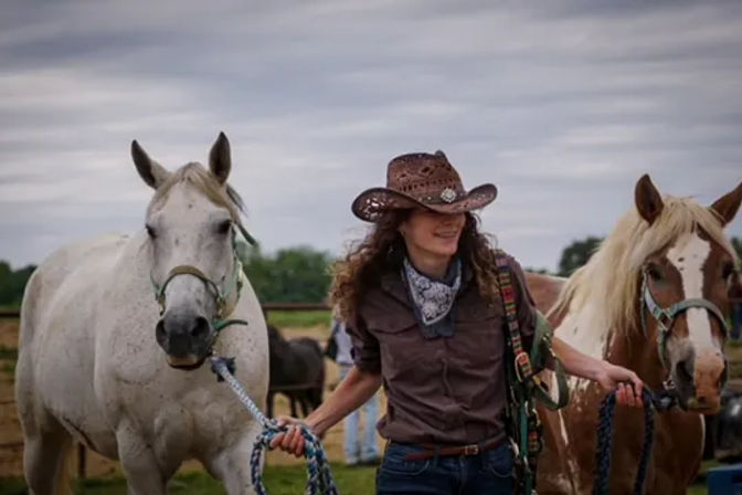 Cowgirl in a brown cowboy hat and bandana leading a white horse and a brown-and-white horse across a grassy ranch pasture under a cloudy sky.