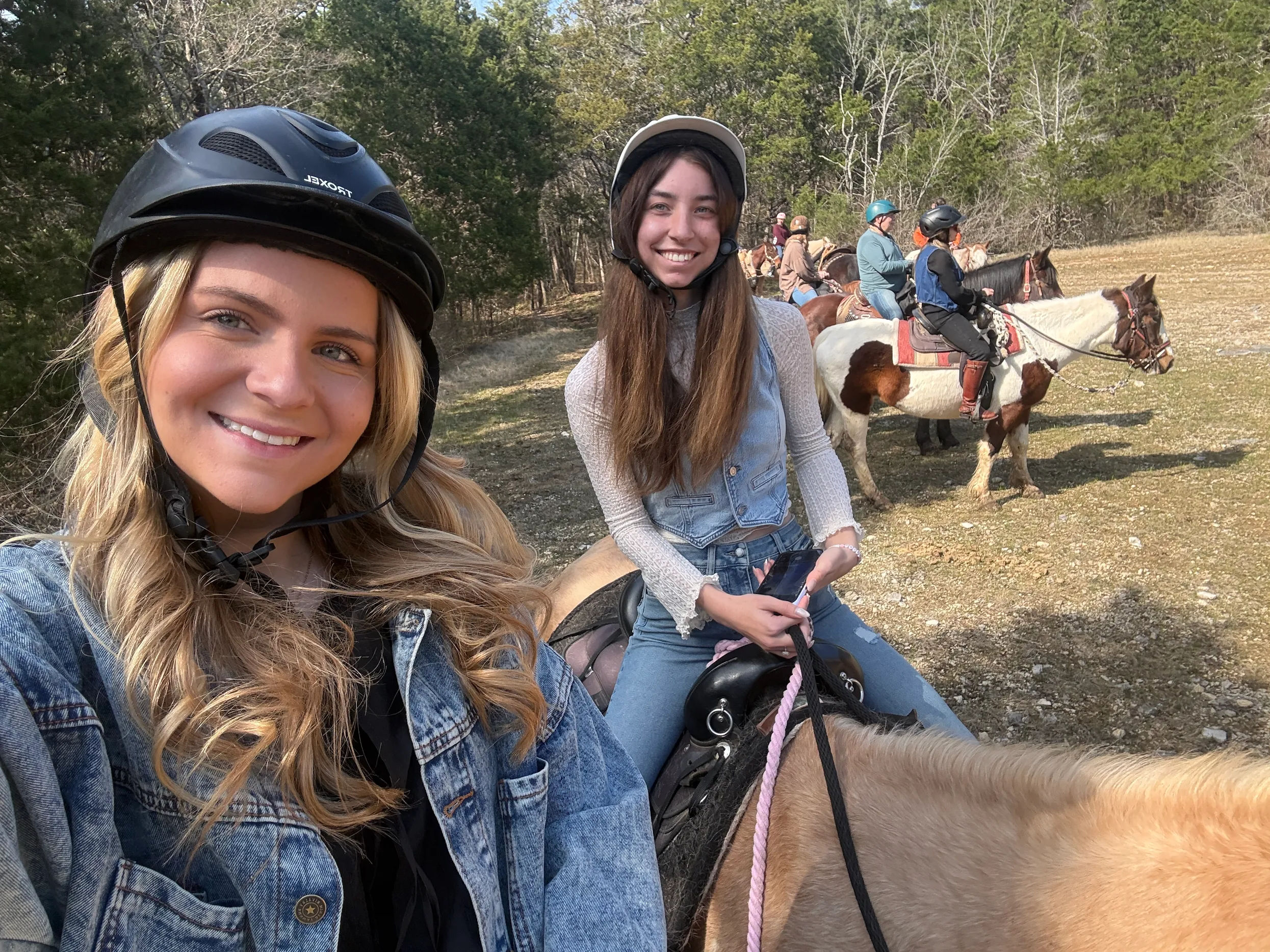 Selfie of two smiling women wearing riding helmets on a sunny horseback trail in a wooded field, one on a light tan horse and the other holding a phone, with additional riders and a pinto horse in the background.