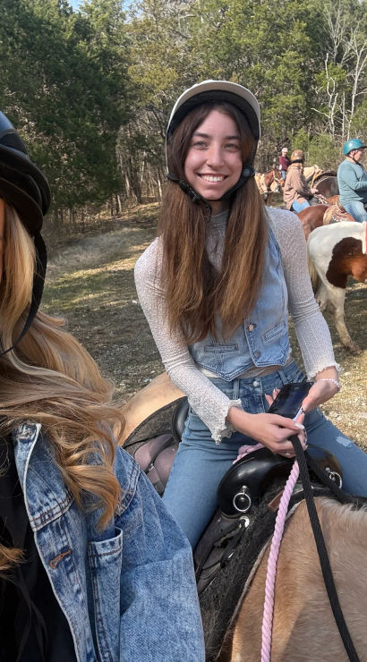 Selfie of two smiling women wearing riding helmets on a sunny horseback trail in a wooded field, one on a light tan horse and the other holding a phone, with additional riders and a pinto horse in the background.