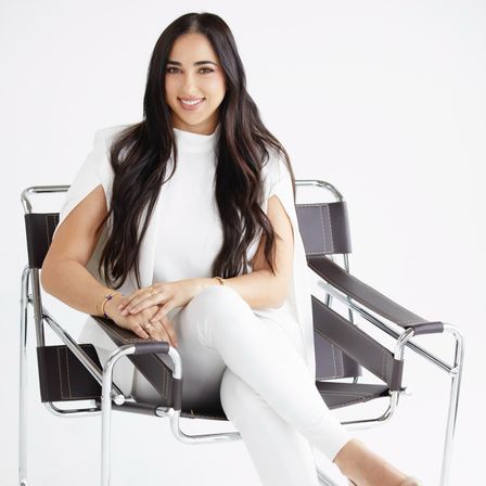 Smiling woman with long dark hair in an all-white outfit seated cross-legged in a modern chrome-and-leather chair on a white studio background, hands folded and wearing bracelets and rings.