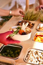 Gloved hand placing a wooden grazing bowl with cheese wedges, olives, dried apricots, cherries, crackers and rosemary at a self-serve snack bar with stainless steel bins of olives and pistachios