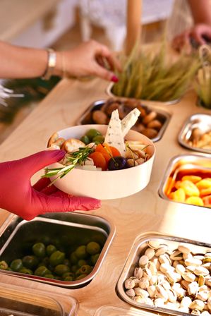 Gloved hand placing a wooden grazing bowl with cheese wedges, olives, dried apricots, cherries, crackers and rosemary at a self-serve snack bar with stainless steel bins of olives and pistachios