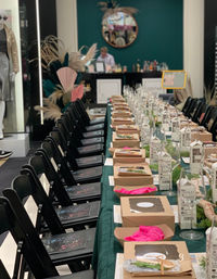 Banquet-style pop-up dinner setup inside a retail store: long green-table runner lined with boxed meals, wine glasses, boxed water, black folding chairs, pampas decor and round mirror in background.