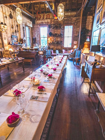 Long rustic barn dining room with a banquet table set with glassware, pink rose petals, vintage farmhouse decor and warm ambient lighting.