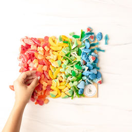 Hand reaching for a rainbow of sugar-coated gummy candies arranged red to blue on a white marble background