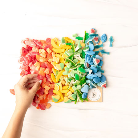 Hand reaching for a rainbow of sugar-coated gummy candies arranged red to blue on a white marble background