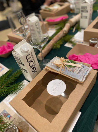Festive workshop tablescape: kraft gift box with clear window, water carton and wine glass, bundled wooden utensils, pink tissue and evergreen sprigs on a green tablecloth.